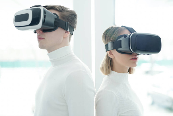 Young people in white sweaters wearing virtual reality goggles standing back to back - Photo, Image