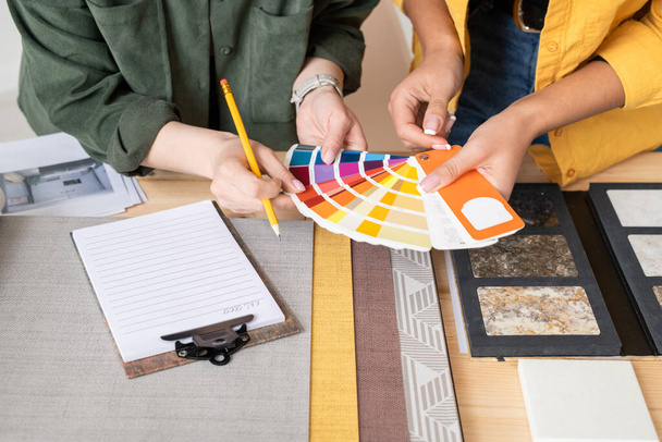Руки двух молодых женщин-дизайнеров интерьеров консультируют о выборе цвета для одной из комнат, держа палитру над столом
 - Фото, изображение