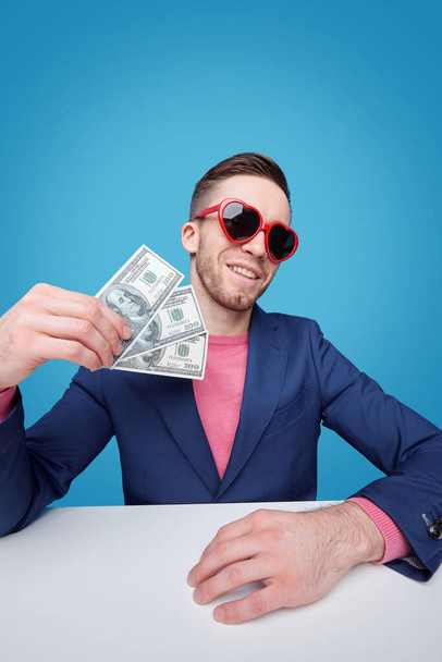 Elegante joven rico en chaqueta azul y gafas de sol glamorosas con billetes de tres dólares mientras está sentado junto al escritorio
 - Foto, imagen