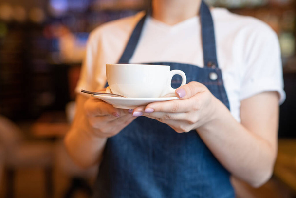 Young waitress in blue apron holding or carrying cup of tea or cappuccino on saucer for one of clients of cafe or restaurant - Фото, изображение