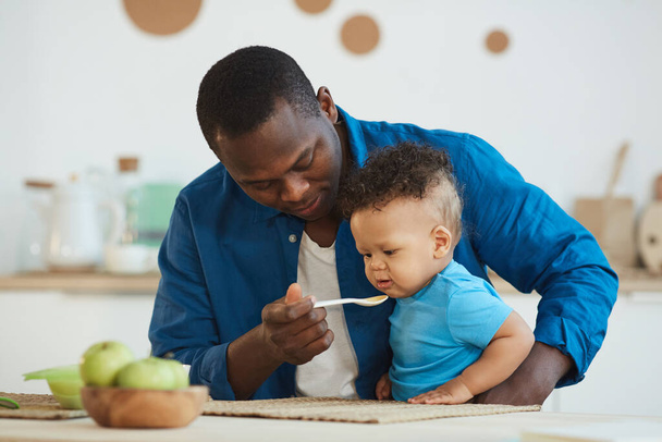 Retrato de feliz pai afro-americano alimentando bonito menino branco sentado na mesa da cozinha
 - Foto, Imagem