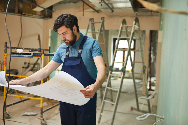 Portrét vousatého stavebního dělníka při pohledu na půdorysy při samotné renovaci domu, kopírovací prostor - Fotografie, Obrázek