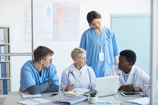 Многонациональная группа врачей, общающихся за столом с ноутбуком в интерьере клиники, копировальное пространство
 - Фото, изображение