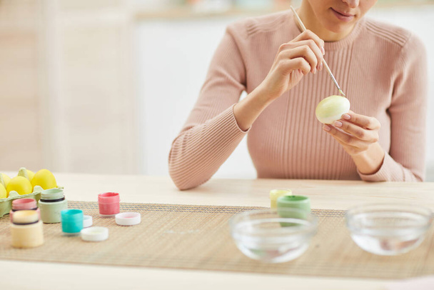 Retrato recortado de mujer adulta sonriente pintando huevos de Pascua mientras está sentada en la mesa en el acogedor interior de la cocina, espacio para copiar
 - Foto, imagen