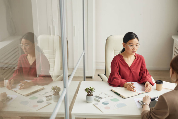 Άποψη υψηλής γωνίας στη σύγχρονη ασιατική επιχειρηματίας μιλώντας με τον πελάτη, ενώ εργάζονται στο γραφείο σε λευκό γραφείο cubicle, αντίγραφο χώρο - Φωτογραφία, εικόνα