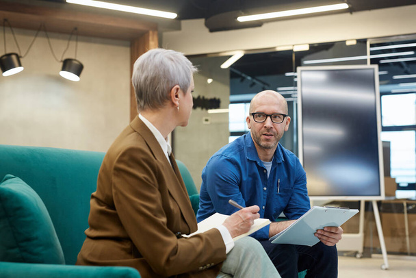 Portrait of bald mature man discussing work project with female manager while sitting on couch in modern office interior, copy space - Photo, Image