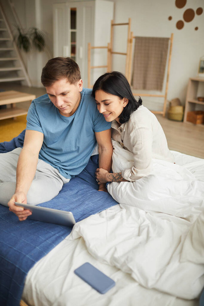 Vertical high angle portrait of young happy couple using digital tablet while sitting on bed at home and ordering food online or watching movie, copy space - Zdjęcie, obraz