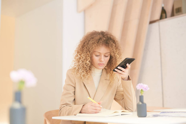 Teplo-tónovaný portrét kudrnaté-vlasy mladá žena pomocí smartphonu při práci nebo studiu posezení u stolu v kavárně, kopírovací prostor - Fotografie, Obrázek