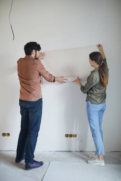 Full length back view portrait of married couple holding wallpaper to wall while redecorating house, copy space - Foto, Bild