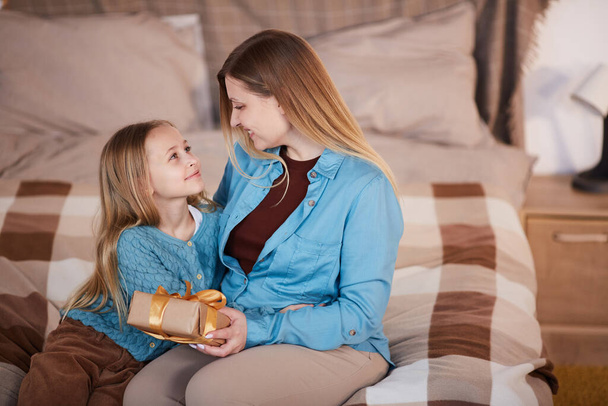 High angle portrait of happy mother hugging cute girl and smiling at each other while opening present sitting on bed, copy space - Photo, Image