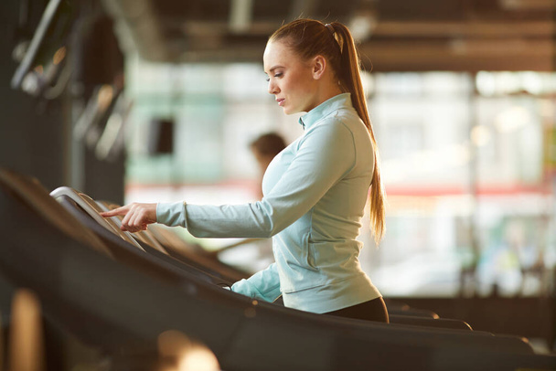 Seitenansicht Porträt einer sportlichen jungen Frau, die im sonnenbeschienenen Fitnessstudio ein Laufband für Cardio-Workout aufbaut, Kopierraum - Foto, Bild