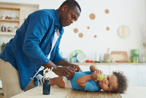 Side view portrait of African-American father changing diaper to cute African-American toddler eating apple, copy space - Foto, imagen