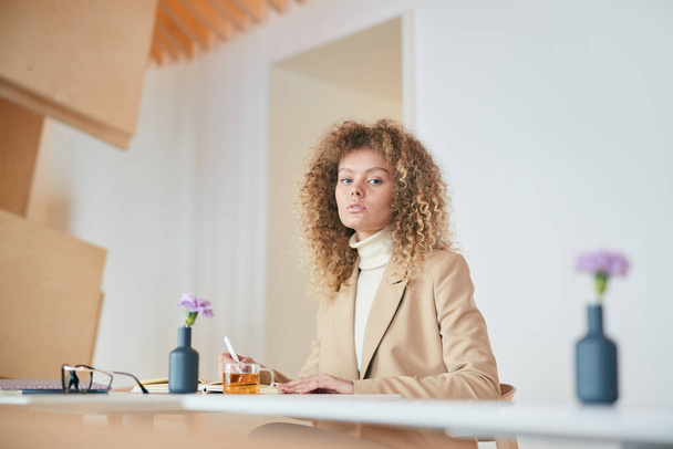 Weitwinkelporträt einer lockigen jungen Frau, die während ihrer Arbeit oder ihres Studiums in die Kamera schaut, am Tisch im Café sitzend, Kopierraum - Foto, Bild