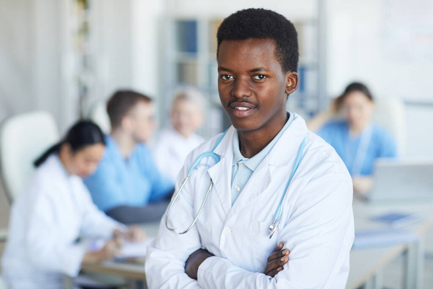 Waist up portrait of young African-American doctor standing with arms crossed and smiling at camera against medical conference background, copy space - Fotó, kép