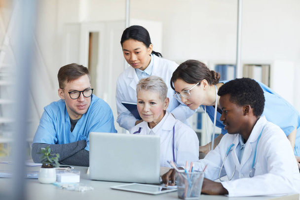 Multietnická skupina lékařů, kteří společně používají notebook, zatímco sedí za stolem v interiéru lékařské ordinace - Fotografie, Obrázek