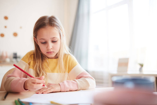 Warm toned portrait of cute little girl drawing pictures or doing homework while sitting at table in home interior, copy space - Photo, Image