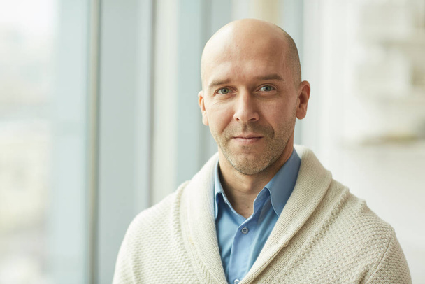 Head and shoulders portrait of balding mature man wearing cardigan looking at camera while standing by window in white office, copy space - Photo, Image