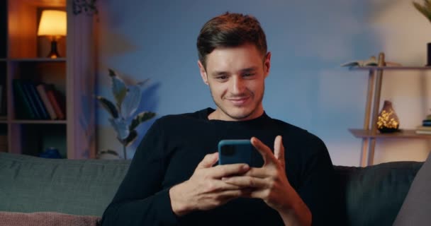 Cheerful millennial man communicating with friends in social networks and laughing while getting funny message. Handsome young guy looking at screen and typing while sitting on sofa. - Footage, Video