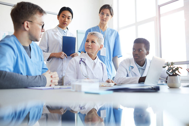 Orvosok soknemzetiségű csoportja, akik együtt dolgoznak az orvosi rendelő belsejében ülve, reflektálással fénymásolt térben. - Fotó, kép