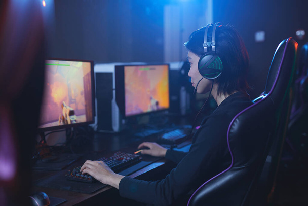 Visão lateral retrato de jovem asiático jogando videogames e usando fones de ouvido no interior cibernético escuro, espaço de cópia
 - Foto, Imagem