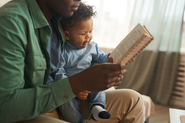 Vista lateral de cerca de cuidar a papá afroamericano leyendo libro a lindo hijo bebé en casa, espacio de copia
 - Foto, Imagen