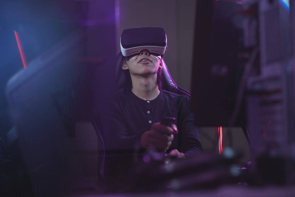 Άποψη χαμηλής γωνίας σε νεαρό Ασιάτη που φοράει ακουστικά VR ενώ παίζει βιντεοπαιχνίδια χρησιμοποιώντας αγωνιστική μετατόπιση στο σκοτεινό εσωτερικό του κυβερνοχώρου, αντιγραφή χώρου - Φωτογραφία, εικόνα