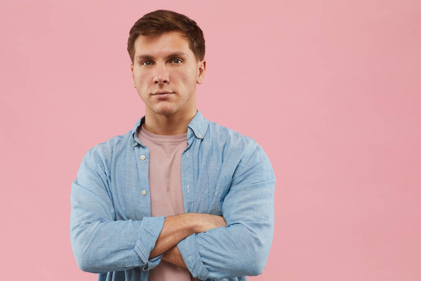 Cintura hacia arriba retrato de un hombre serio de pie con los brazos cruzados y mirando a la cámara mientras posa sobre fondo rosa en el estudio, espacio de copia
 - Foto, imagen