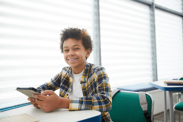 Retrato de un alegre niño afroamericano que tiene teléfono inteligente mientras se sienta en el escritorio en el aula escolar y mira la cámara, copie el espacio. - Foto, imagen