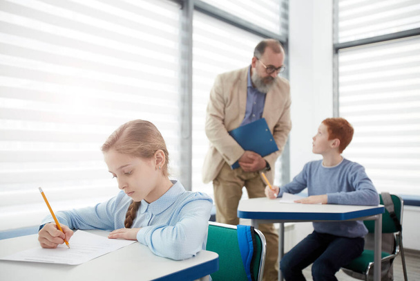 Group of children taking test with bearded mature teacher watching them, focus on cute blond girl sitting at desk and writing in foreground, copy space - Fotó, kép