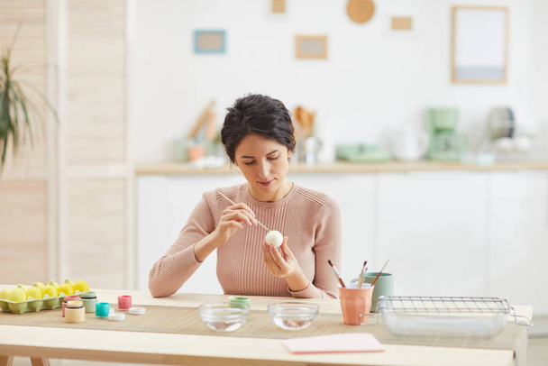 Портрет сучасної дорослої жінки, що малює великодні яйця, сидячи за столом у затишному інтер'єрі кухні, копіює простір
 - Фото, зображення