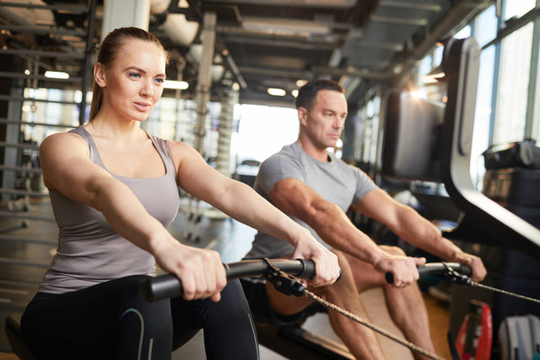 Portrait of sportive couple using exercise machines during strength workout in modern gym, copy space - 写真・画像