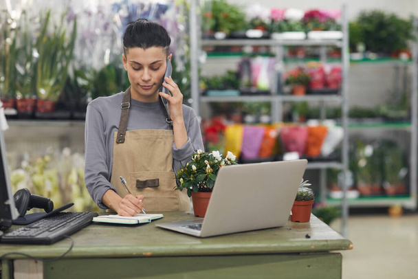 Waist up portrait of female small business owner speaking by smartphone while standing behind counter in flower shop, copy space - Foto, afbeelding
