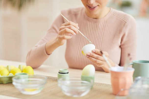 Retrato recortado de una joven sonriente pintando huevos de Pascua mientras está sentada en la mesa en el acogedor interior de la cocina, espacio para copiar
 - Foto, Imagen