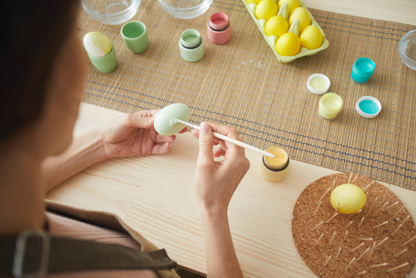 Hohe Nahaufnahme einer unkenntlichen jungen Frau, die Eier in Pastellfarben für Ostern bemalt, während sie am Tisch in Küche oder Kunstatelier sitzt, Kopierraum - Foto, Bild