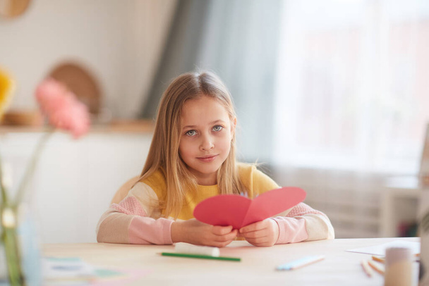 Portrait of cute little girl holding heart-shaped card and looking at camera while sitting at table in cozy home interior, copy space - Fotografie, Obrázek