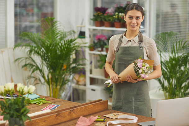 Waist up portrait of young female florist holding beautiful Spring bouquet in flower shop and smiling at camera, copy space - Photo, image