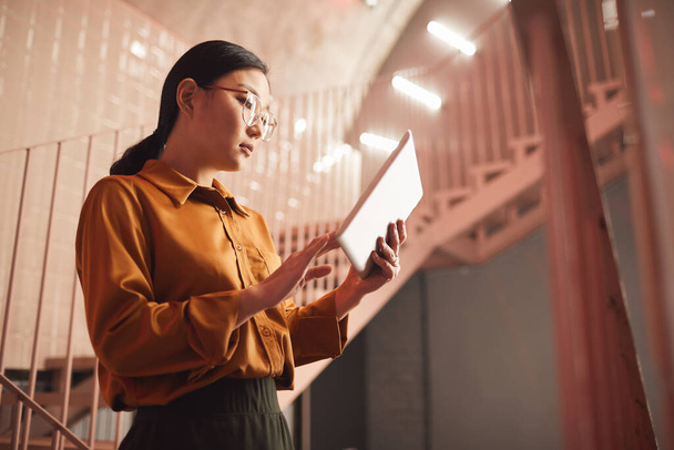 Side view portrait of young Asian businesswoman using tablet while standing by stairs outdoors, copy space - Φωτογραφία, εικόνα