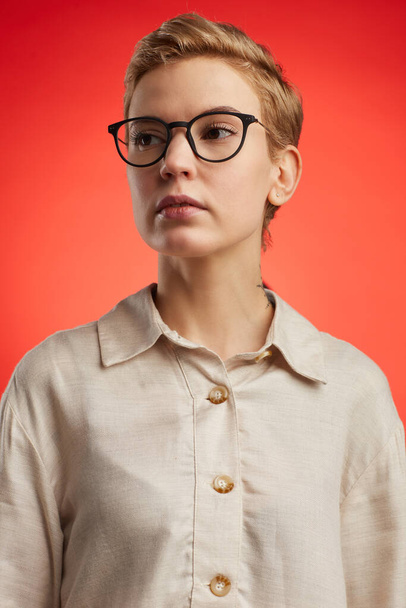 Portrait of modern woman wearing black rimmed glasses with short pixie haircut standing against fiery red background in studio - Photo, Image