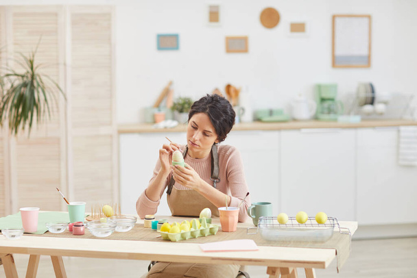 Wide angle view at elegant adult woman painting eggs in pastel colors for Easter while sitting at table in kitchen or art studio, copy space - Fotó, kép
