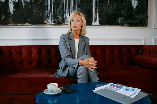 Full length portret van moderne volwassen zakenvrouw op zoek naar camera terwijl zitten op fluwelen bank in cafe, kopieer ruimte - Foto, afbeelding