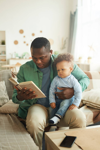 Κάθετη πορτρέτο του ώριμου Αφροαμερικανού διαβάζοντας βιβλίο για το παιδί κάθεται στην αγκαλιά του πατέρα - Φωτογραφία, εικόνα