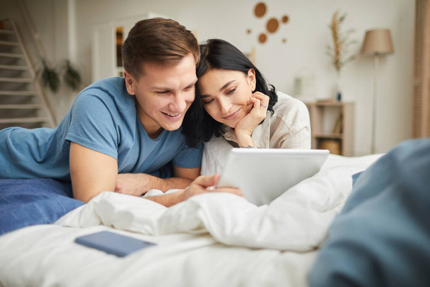 Portrait de jeune couple heureux utilisant une tablette numérique tout en étant couché sur le lit à la maison et en commandant de la nourriture en ligne ou en regardant un film, copiez l'espace
 - Photo, image