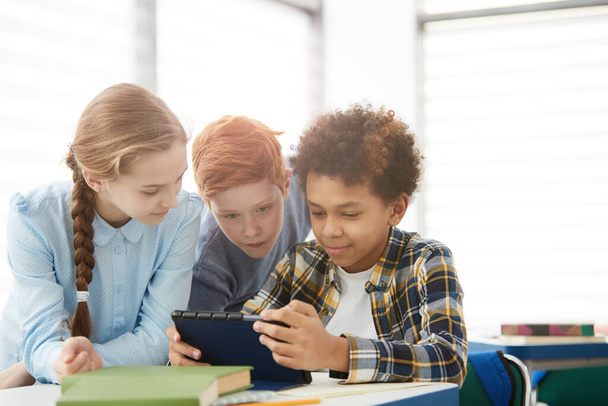 Digitális táblagépet vagy internetet használó gyermekek többnemzetiségű csoportja, akik együtt ülnek az íróasztalnál az osztályteremben - Fotó, kép