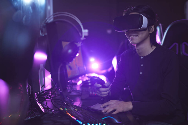 Πλάγια άποψη πορτρέτο του νεαρού Ασιάτη άνδρα που φοράει VR ακουστικά ενώ παίζει βιντεοπαιχνίδια χρησιμοποιώντας αγωνιστικά shift στο σκοτεινό εσωτερικό του κυβερνοχώρου, αντίγραφο χώρου - Φωτογραφία, εικόνα