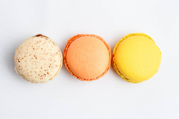 Κάτοψη σειράς τριών γλυκών γαλλικών μακάρων με διαφορετικά αρώματα, που απομονώνονται σε λευκό, γεύσεις λεμονιού, βανίλιας και πορτοκαλιού, νόστιμο επιδόρπιο σε παστέλ χρώματα σε ένα τραπέζι - Φωτογραφία, εικόνα