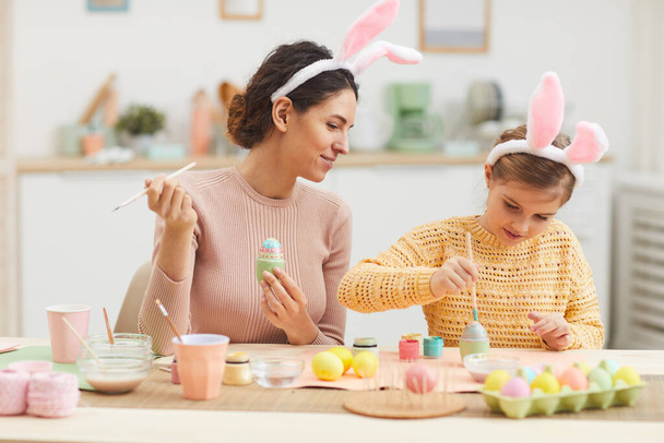 Πορτρέτο της νεαρής μητέρας με το κοριτσάκι που διασκεδάζει ενώ ζωγραφίζει πασχαλινά αυγά στο ζεστό εσωτερικό της κουζίνας, και οι δύο φορώντας αυτιά λαγουδάκι, αντίγραφο χώρου - Φωτογραφία, εικόνα