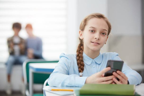 Retrato de la chica rubia linda que tiene teléfono inteligente mientras se sienta en el escritorio en el aula escolar y mira a la cámara, copia el espacio. - Foto, imagen