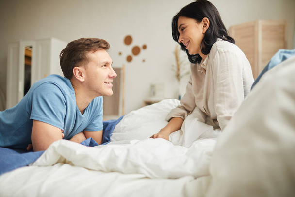 Side view portrait of happy young couple looking at each other while sitting on bed in morning, copy space - Photo, Image