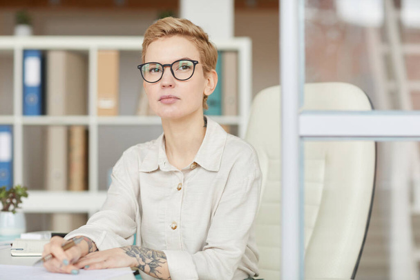 Portrait of confident tattoed businesswoman looking at camera while sitting at desk in office, copy space - Photo, Image