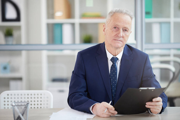 Portret odnoszącego sukcesy starszego biznesmena trzymającego schowek podczas pozowania przy biurku w biurze, przestrzeń do kopiowania - Zdjęcie, obraz